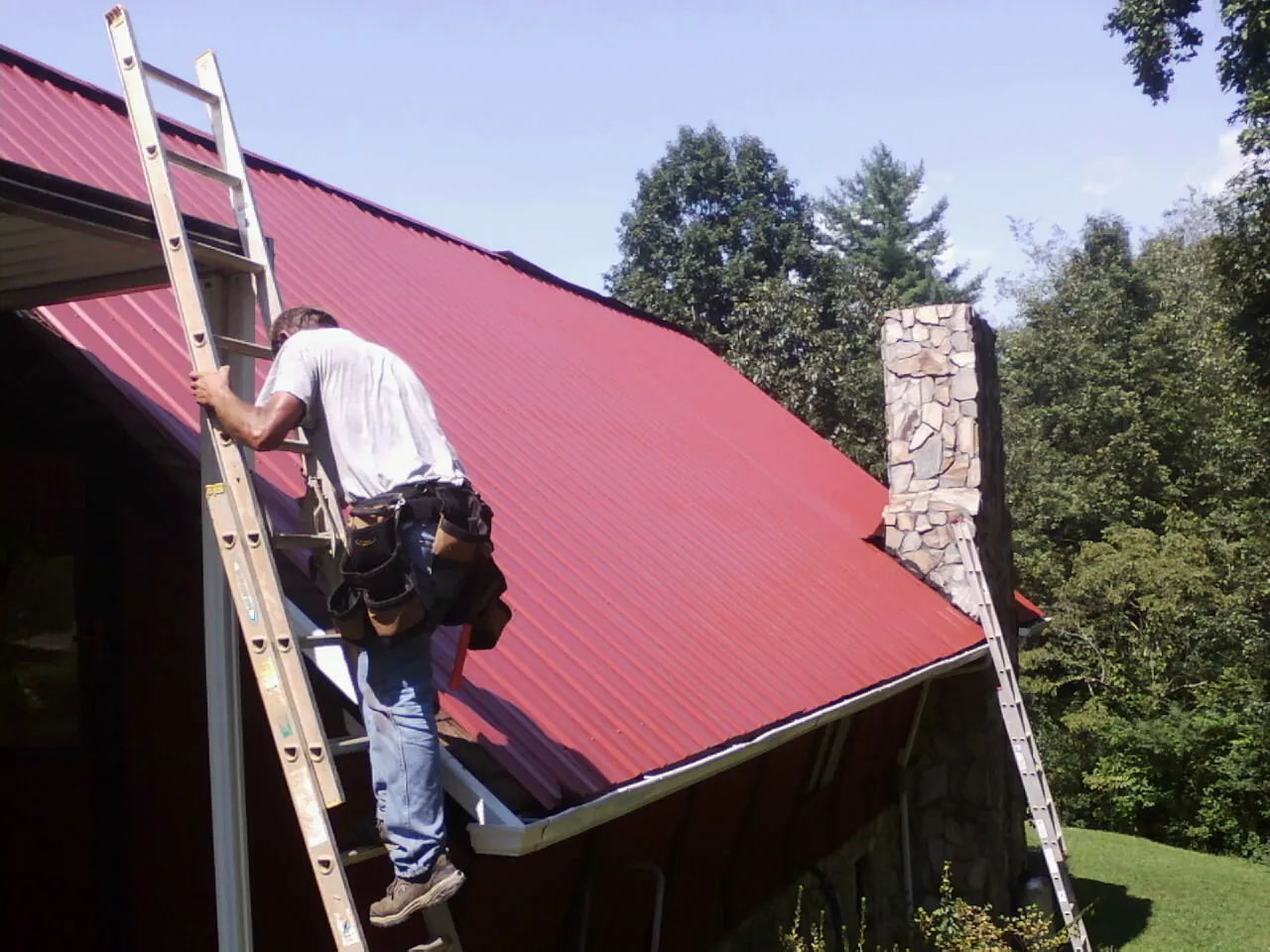 Roofer on a ladder beside a red metal roof and stone chimney — Soffit & Fascia Repair by Roofing Brothers in West Point